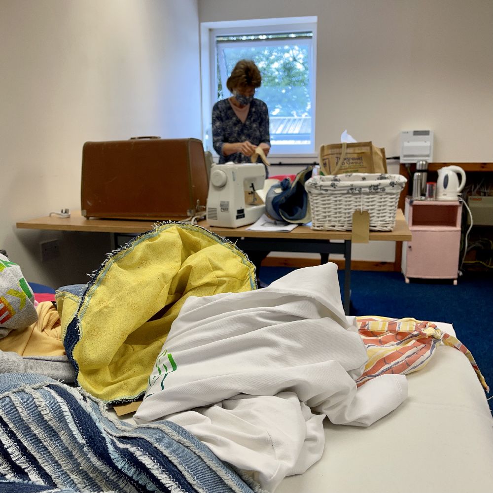 Photo of clothes ready for upcycling in the foreground, with a volunteer working beside a sewing machine in the background
